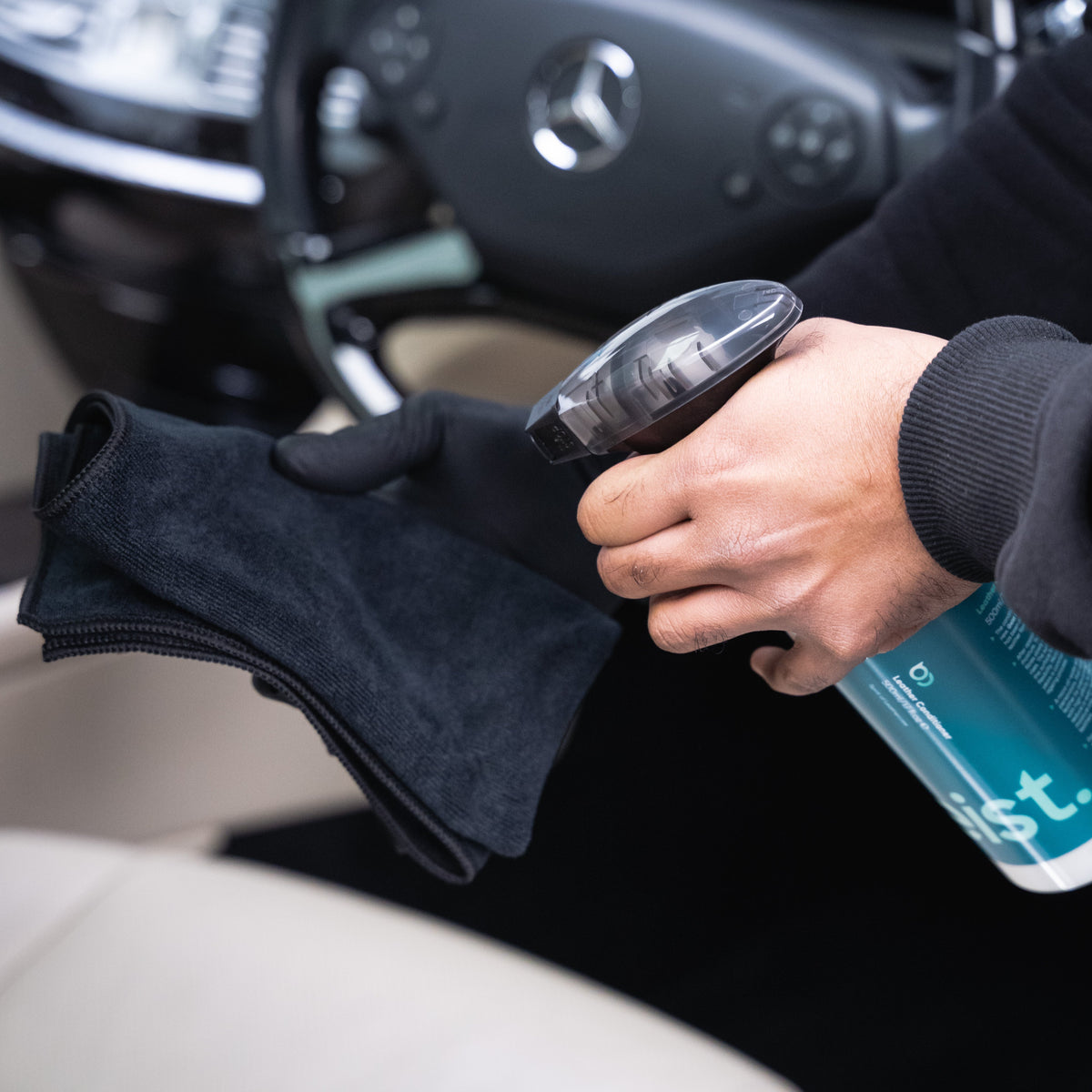 Person cleaning car interior with a spray bottle and black cloth, Mercedes-Benz logo visible.