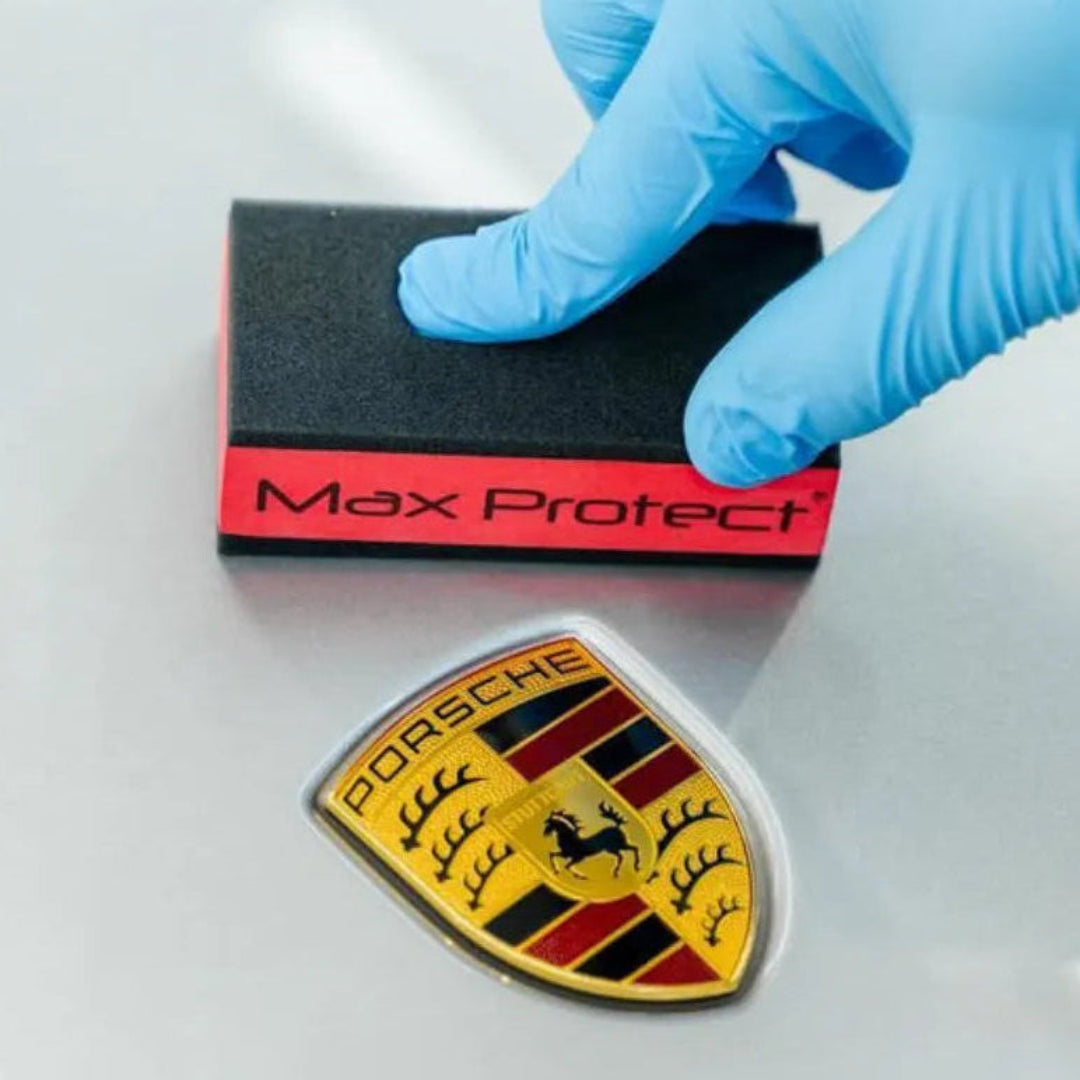 Person wearing blue gloves applying Max Protect product to a Porsche emblem.