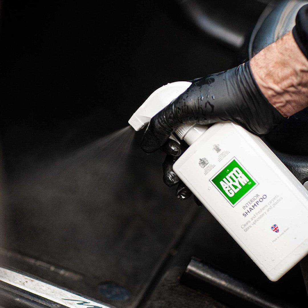 The Detail Warehouse Autoglym Interior Shampoo being sprayed onto a car floor mat by a person wearing black gloves.