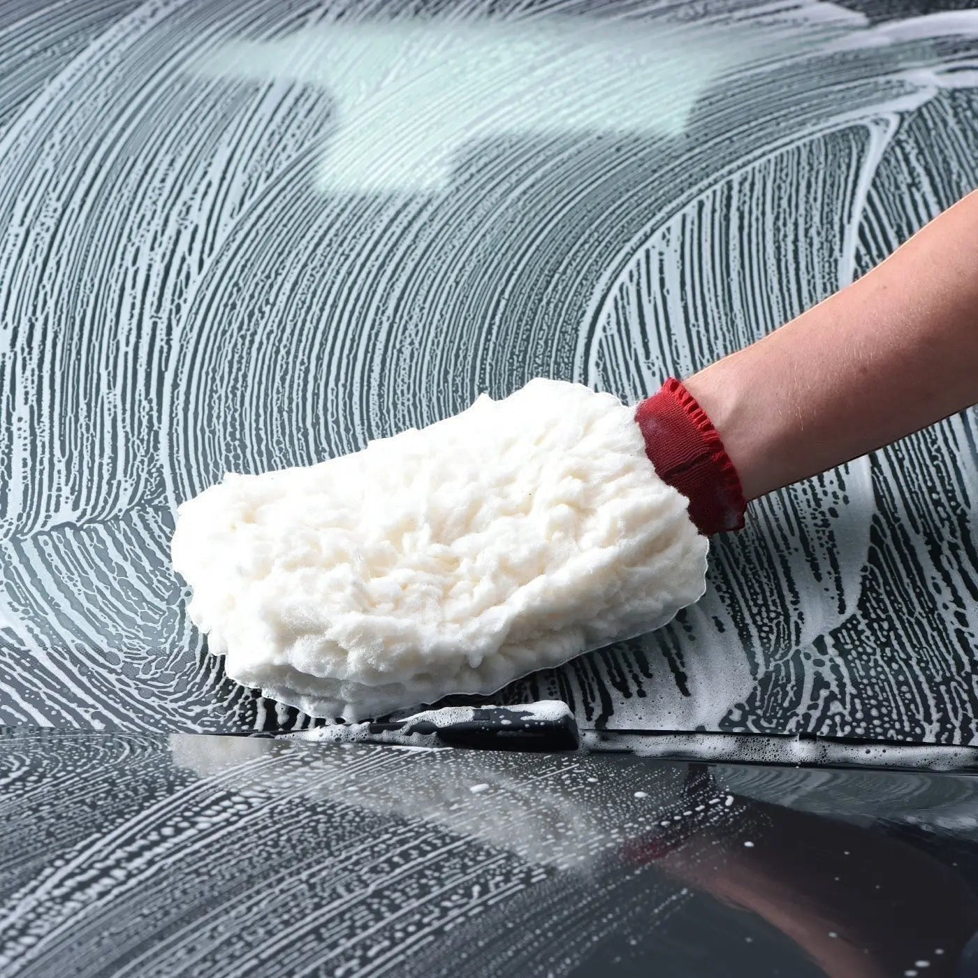 The Detail Warehouse hand using a white microfiber wash mitt to clean a car hood covered in thick, foamy car shampoo.